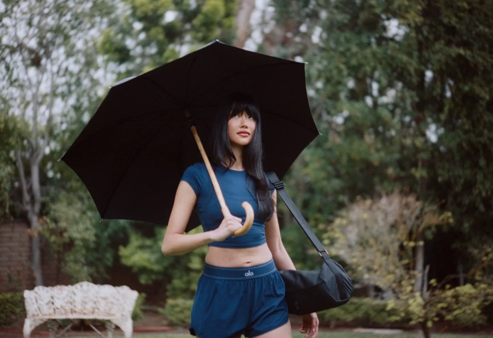 A woman in workout clothes holding an umbrella, gym bag over her shoulder